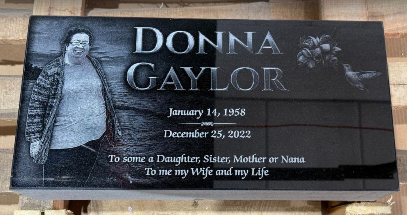 Flat marker for Donna Gaylor — laser-engraved portrait with hummingbird motif and waves and sunset background scene on polished black granite