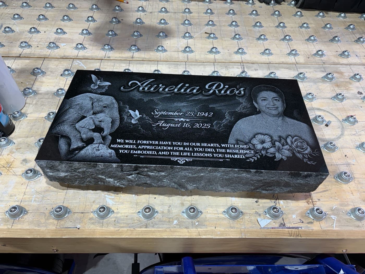 Black granite flat marker for Aurelia Rios — laser-engraved portrait with elephant motif and dove imagery, photographed on the production workbench