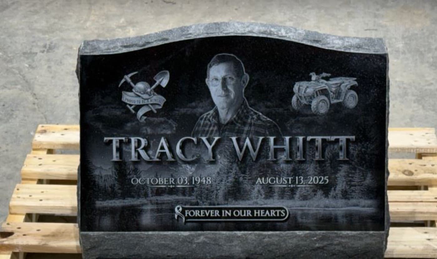 Black granite slant headstone for Tracy Whitt — laser-engraved portrait with miner's emblem, ATV icon, and mountain forest scene on the steep angled face